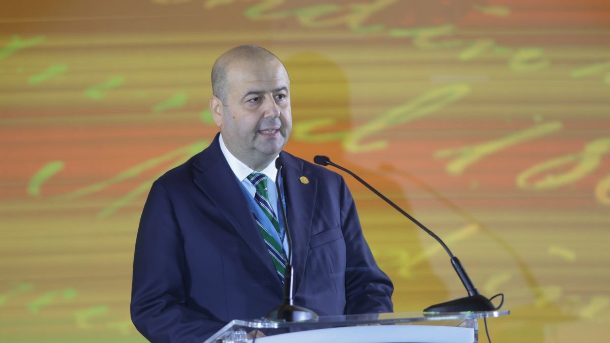 Mihai Busuioc, in costum inchis la culoare si cravata in dungi nuante de albastru si bleu, in timp ce tine un discurs la un eveniment aniversar, in spate un ecran mare in nuante de galben si portocaliu, septembrie 2024