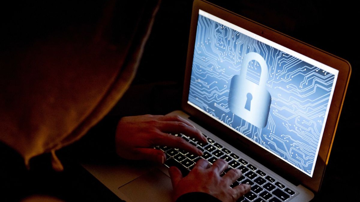 Un bărbat cu glugă care seamănă cu un hacker lucrează la un laptop care are afișat pe ecran un lacăt pe funal albastru deschis.