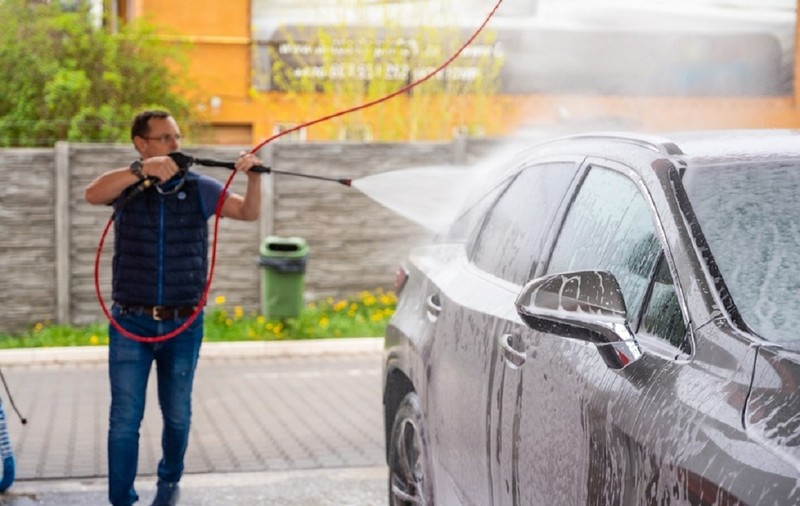Greșeala pe care o fac șoferii când spală mașina la spălătoriile Self-Wash - Un bărbat clătește un autoturism  gri cu jet de apă sub presiune la o spălătorie 