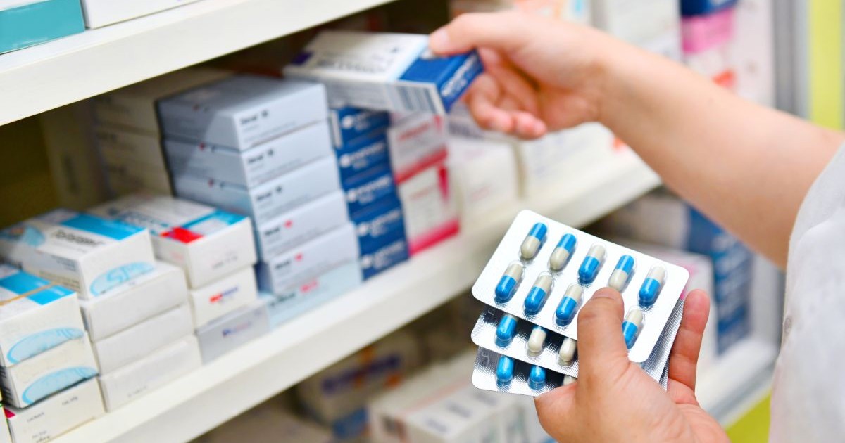 O farmacistă ține în mână o cutie cu medicamente pe care o pune pe raftul farmaciei, iar în mâna stângă are câteva blistere cu pastile