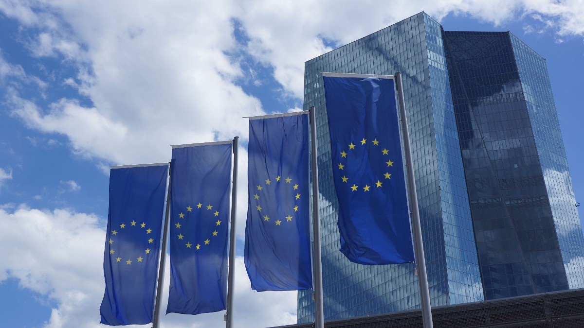 Patru steaguri ale Uniunii Europene flutură pe catarge înalte în fața unei clădiri moderne, compuse din turnuri acoperite cu sticlă de culoare închisă. Steagurile de culoare albastru intens, cu cercul de stele aurii, se profilează pe un fundal de cer albastru cu nori albi pufoși.