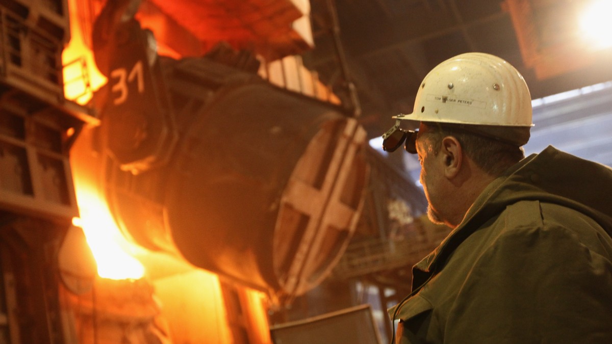 Imaginea prezintă un muncitor cu cască de protecție care supraveghează un recipient masiv industrial în timpul turnării sau prelucrării metalului topit într-o oțelărie.