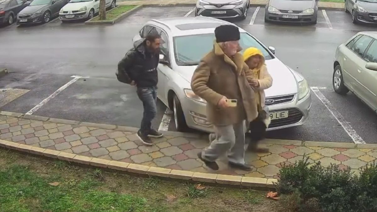 Imaginea surprinde o femeie îmbrăcată într-o haină deschisă la culoare, cu o glugă galbenă pe cap ținând mai multe sacoșe în mâini, un bărbat cu părul alb, îmbrăcat cu un cojoc maro și o căciulă neagră pe cap și în spatele lor un bărbat cu părul negru, cu o jachetă neagră, traversând o parcare.