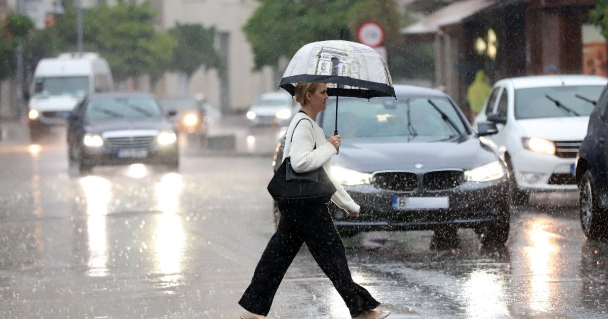 O femeie îmbrăcată cu bluză albă și pantaloni negri traversează strada pe trecerea de pietoni și are în mână o umbrelă deschisă, într-o zi în care plouă, București, martie 2025.