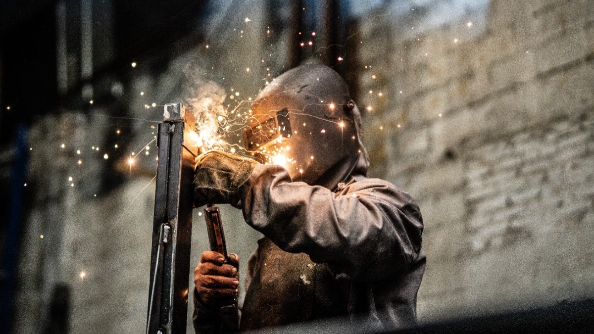 Un sudor purtând mască și echipament de protecție în timp ce sudează o structură metalică, împrăștiind scântei luminoase într-un mediu industrial.