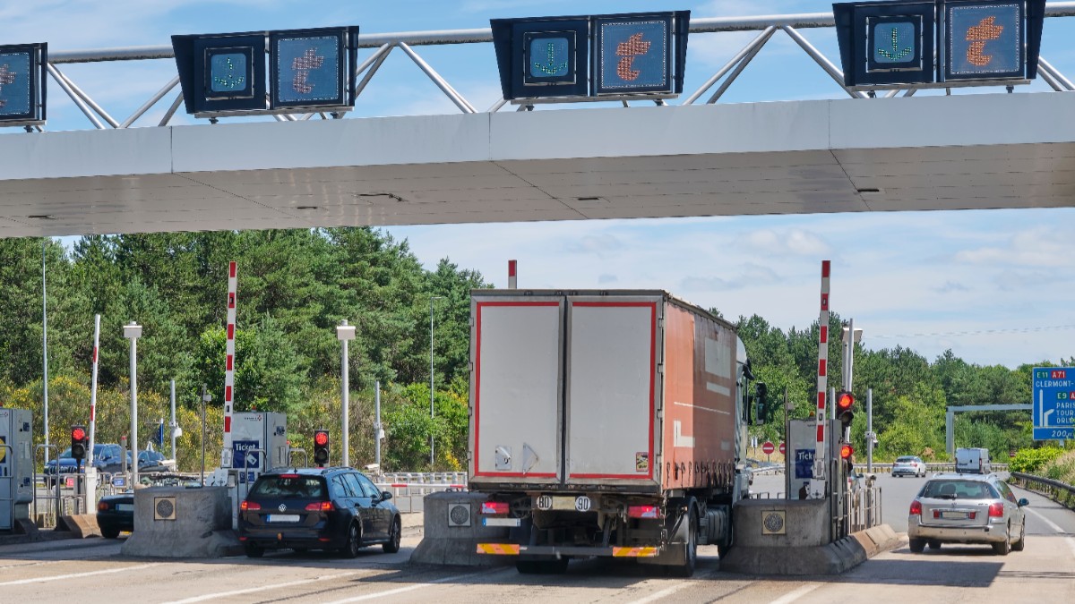 Imaginea înfățișează un camion și câteva autoturisme trecând prin porțile unei stații de taxare de pe o autostradă din Franța.