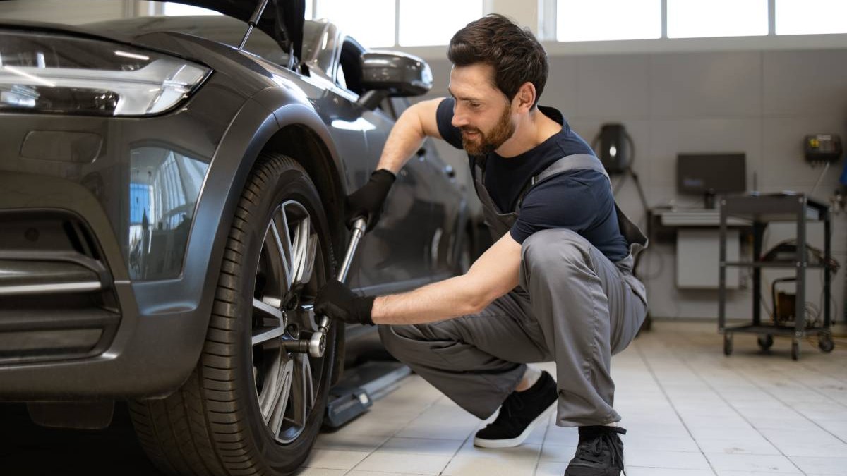 Un mecanic auto îmbrăcat cu salopetă gri și tricou negru, în timp ce strânge prezoanele roții unei mașini negre