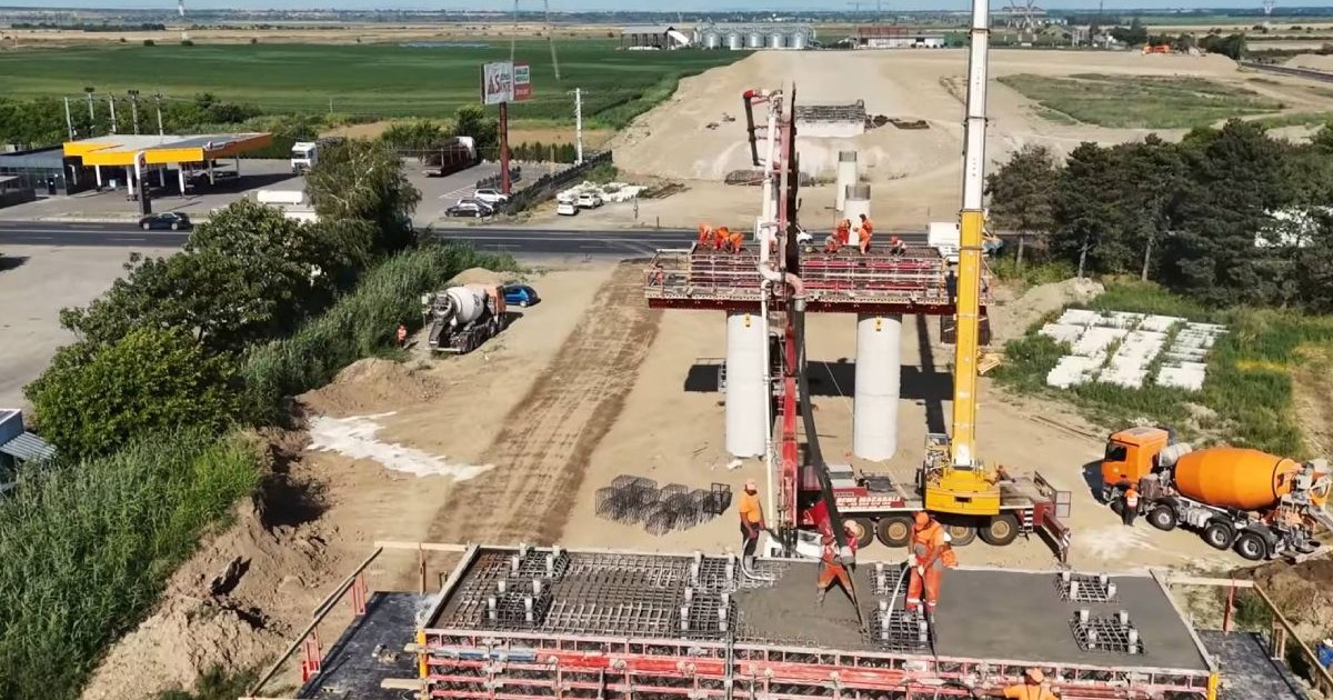 Lucrări la Drum Expres Focsani-Braila-Constanta, in imagini muncitori lucreaza la picioare de pod, se văd utilaje, drumul defrisat, in jurul lui iarba, in departare cerul senin