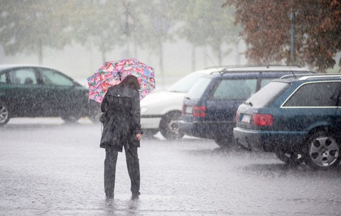 Imaginea prezintă o persoană îmbrăcată în haine închise la culoare care merge sub o umbrelă colorată în timpul unei ploi torențiale, într-o zonă de parcare cu autoturisme.   