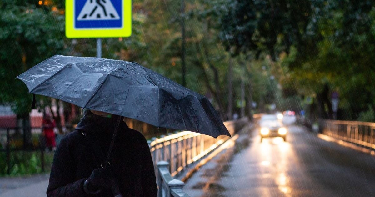 O femeie ține o umbrelă neagră, în timp ce merge pe stradă, iar afară plouă și e frig, iar o mașină cu farurile aprinse vine din față