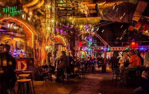 nteriorul unui local aglomerat, amenajat într-un spațiu vast cu un aspect brut, industrial, definit de pereți cu textură nefinisată, arcade ample și stâlpi înalți din schele metalice. Încăperea este decorată abundent și aparent haotic cu o multitudine de obiecte suspendate de tavanul înalt, incluzând globuri disco, lămpi cu abajururi de forme diferite, cabluri la vedere și diverse alte decorațiuni eclectice.