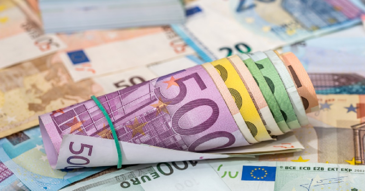 Imaginea prezintă un sul de bancnote euro de diverse cupiuri, fixat cu un elastic, așezat peste o mulțime de alte bancnote împrăștiate, evidențiind valoarea de 500 de euro în prim-plan.