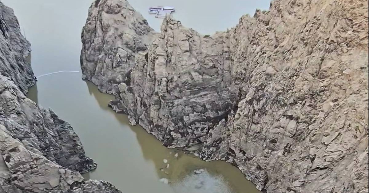 Imagine cu lacul Vidraru, între stânci abrupte. Pe apă se vede o peliculă închisă la culoare, posibil combustibil, iar in departare o alta nava.