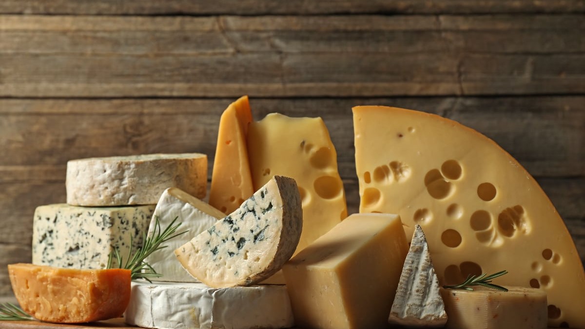 O varietate bogată de brânzeturi este aranjată artistic pe un fundal din lemn rustic, incluzând tipuri precum brânză cu mucegai alb (tip Brie sau Camembert), brânză cu mucegai albastru (tip Roquefort) și felii mari de brânză cu găuri (tip Emmentaler). Brânzeturile sunt așezate pe un tocător din lemn și decorate cu câteva crenguțe de rozmarin, sugerând o degustare sau o planșă de aperitive.