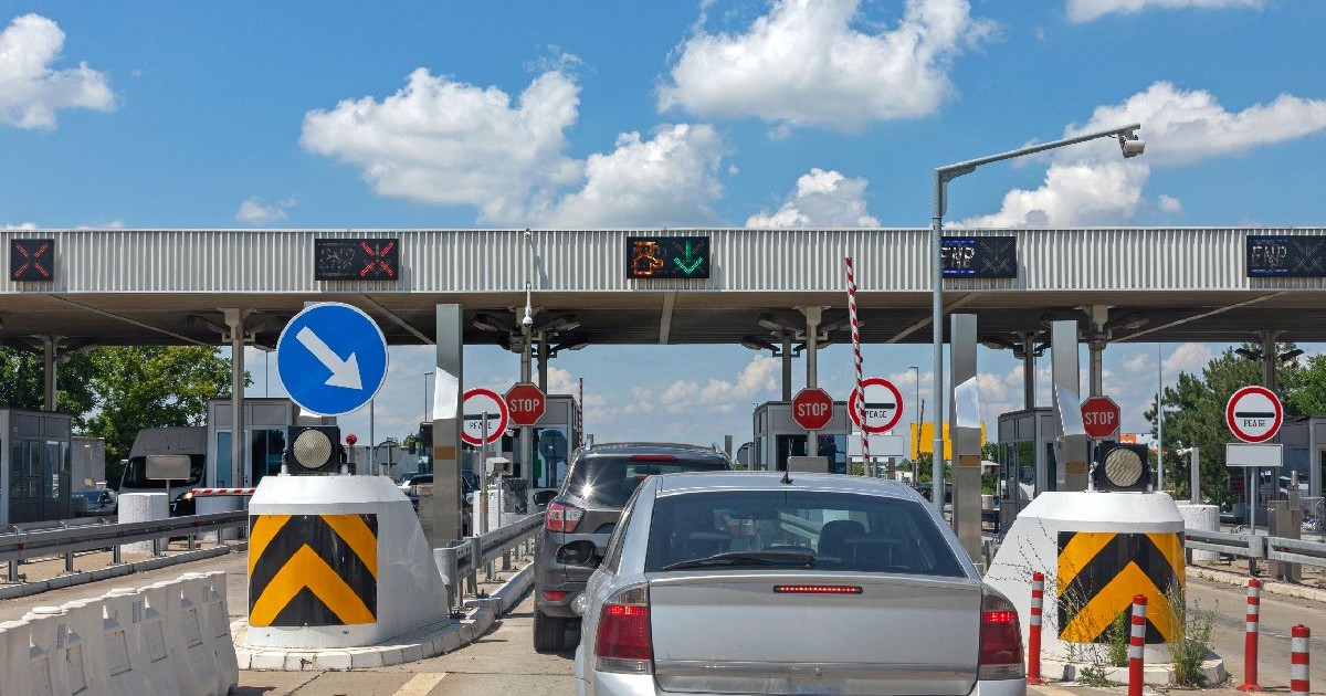Fotografie cu o stație de taxare Sedan argintiu în prim-plan, coadă de mașini, semafoare, indicatoare rutiere și bariere.