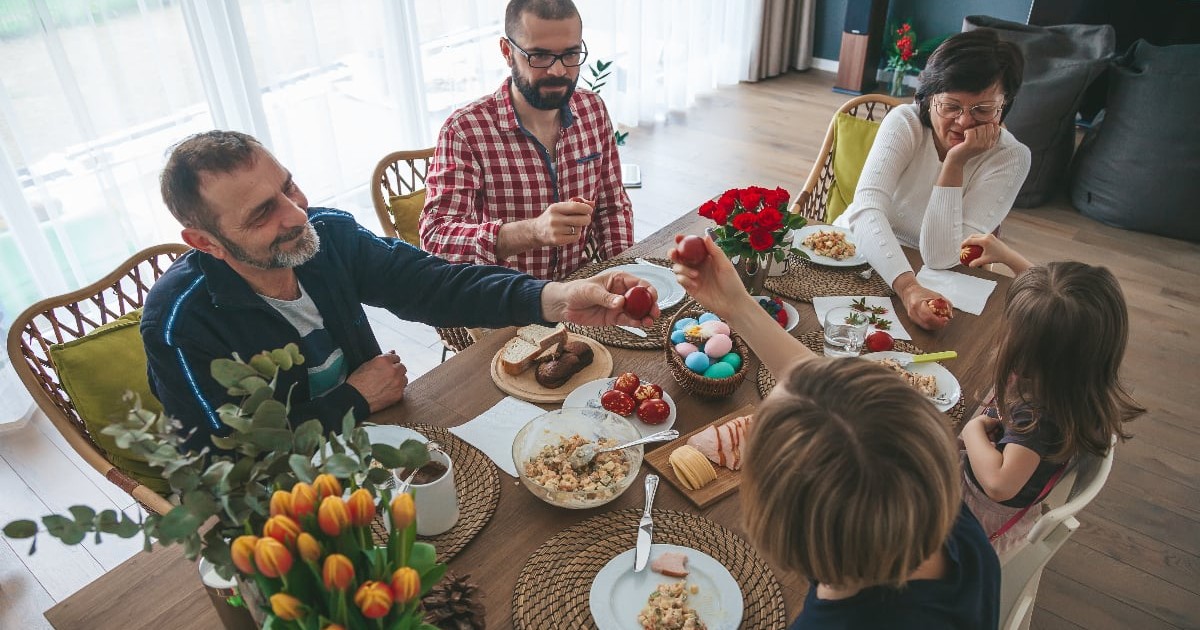 Imaginea surprinde o masă de familie, unde mai multe persoane stau în jurul unei mese, sugerând un moment de reuniune. În fundal se observă un spațiu exterior cu grădini și case, creând o atmosferă de viață cotidiană.
