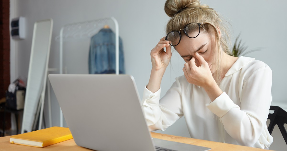 O femeie își ține mâna în jurul ochilor și cu cealaltă ochelarii, după o zi de muncă grea în fața calculatorului