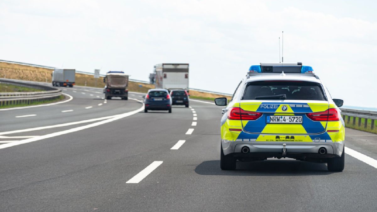 Imaginea arată o autostradă din Germania, având în prim-plan o mașină de poliție în mișcare. În fața autospecialei se află câteva autoturisme și trei camioane.