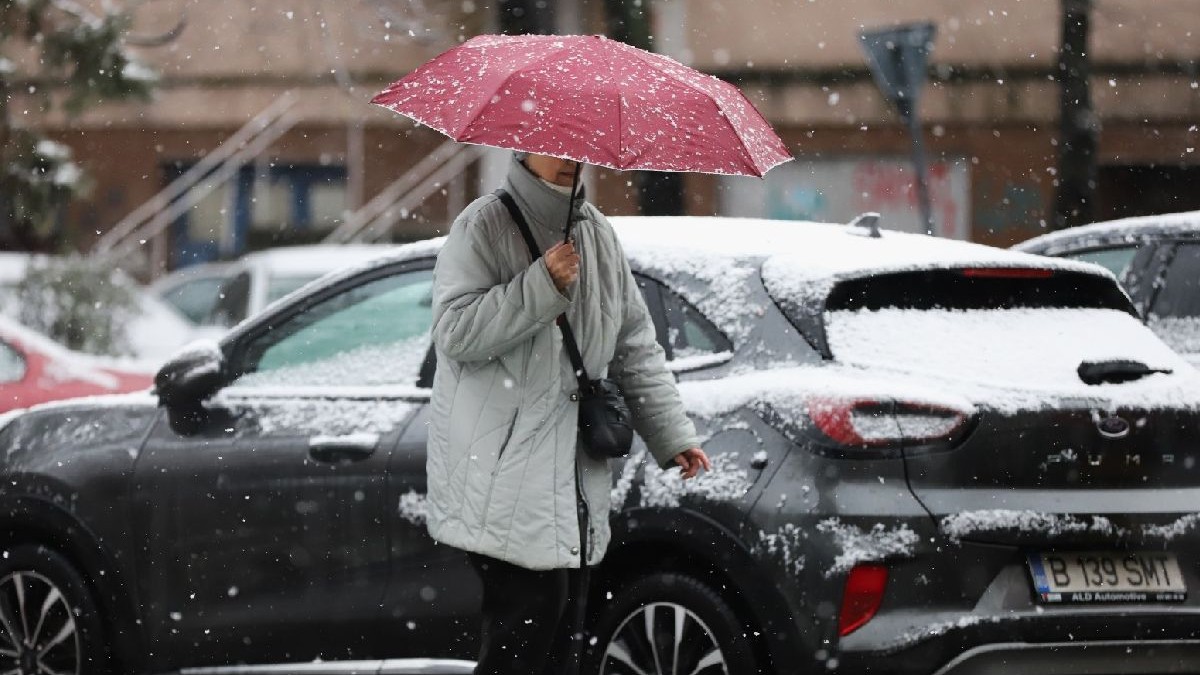 Fotografie cu o femeie cu o umbrelă roșie mergând pe o stradă acoperită de zăpadă, mașini acoperite de zăpadă, clădiri în fundal.
