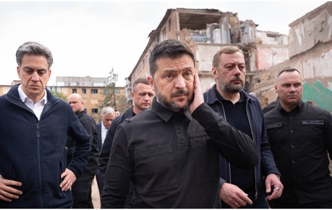 Președintele Ucrainei, Volodimir Zelenski, îmbrăcat în haine închise la culoare de stil militar, mergând alături de oficiali și gărzile sale de corp în fața unei clădiri rezidențiale grav avariate.