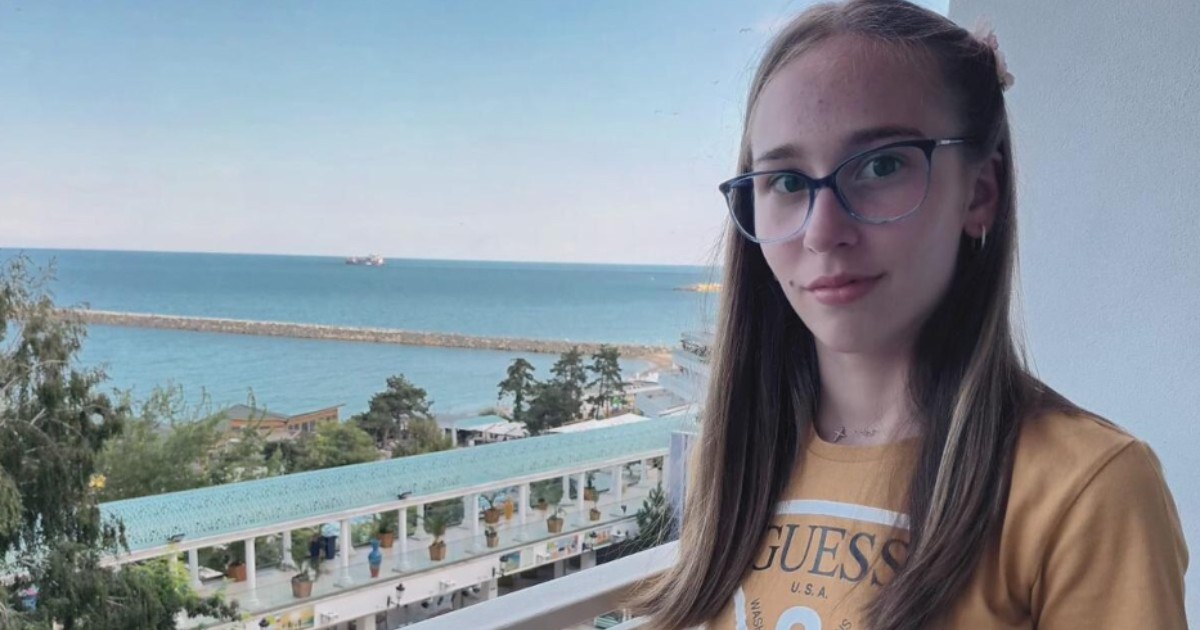 O tânără cu ochelari, care poartă un tricou galben. Aceasta stă pe un balcon, iar în fundal este marea