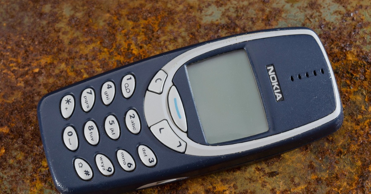 Imaginea prezintă un telefon mobil Nokia 3310, așezat pe o suprafață ruginită, evidențiind designul său clasic și tastatura numerică specifică începutului anilor 2000. Aspectul robust și ecranul monocrom subliniază simplitatea și durabilitatea care au făcut acest model celebru.