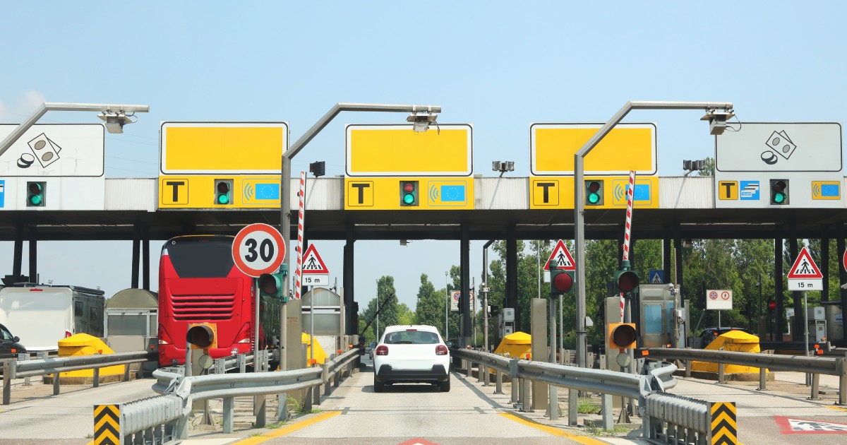 Imaginea prezintă o stație de taxare pe autostradă cu mai multe porți de acces, unde un autoturism alb și un autobuz roșu se pregătesc să treacă prin punctele de control.
