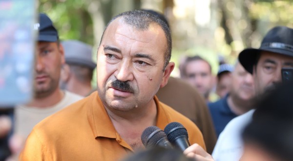 Un bărbat cu mustață vorbind în fața mai multor microfoane, înconjurat de oameni și jurnaliști