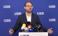 Dominic Fritz îmbrăcat într-un sacou negru și o cămașă albă, vorbind în fața unui pupitru cu sigla USR pe care se află mai multe microfoane, cu mâinile larg deschise