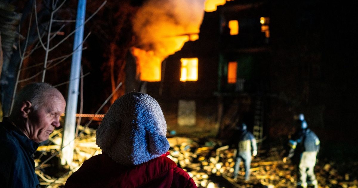 O persoană cu spatele, care poartă o cârpă pe cap, se uită la doi pompieri care încearcă să stingă flăcările la o clădire bombardată în Ucraina, pe timp de noapte