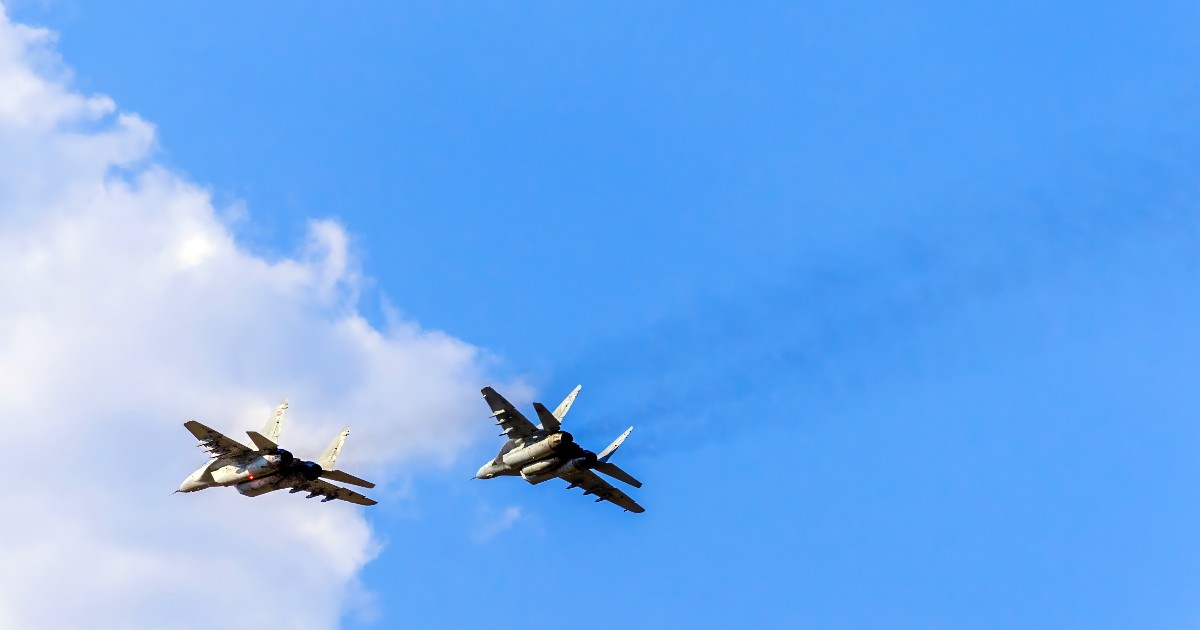 Două avioane de vânătoare zboară în formație strânsă pe un cer predominant albastru, parțial acoperit de câțiva nori albi. Avioanele, văzute de jos, lasă în urmă urme discrete de fum și se deplasează la o viteză mare, probabil executând un zbor demonstrativ sau o misiune.