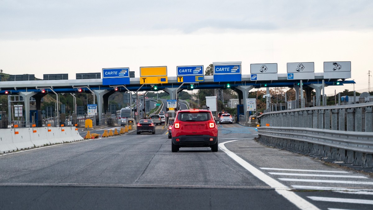Imaginea prezintă mai multe mașini, printre care un SUV roșu, care se apropie de porțile unei stații de taxare de pe o autostradă.
