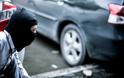 Un bărbat purtând o cagulă neagră, ghemuit discret lângă un autoturism parcat.   