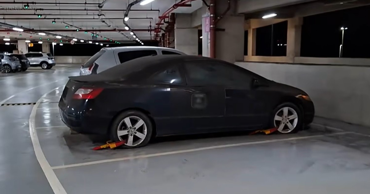 Un autoturism coupe de culoare închisă, acoperit de un strat gros de praf și având ambele roți din imagini blocate cu dispozitive speciale galben-roșii, parcat în interiorul unei parcări subterane sau etajate.