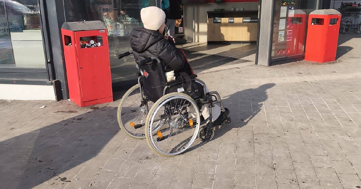 O femeie stă într-un cărucior cu rotile, pe cap poartă un fes