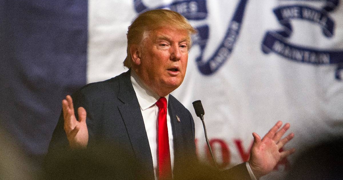 În imagine, Donald Trump ține mâinile larg deschise în timp ce explică ceva la un microfon.