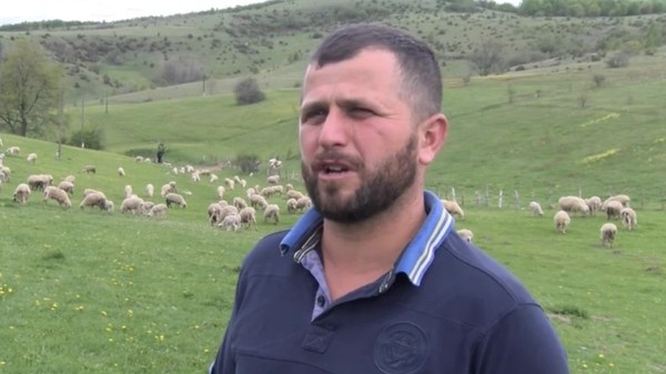 Un bărbat cu barbă, îmbrăcat într-un tricou polo albastru închis, vorbind în aer liber pe o pășune unde paște o turmă de oi, pe fundalul unor dealuri înverzite.
