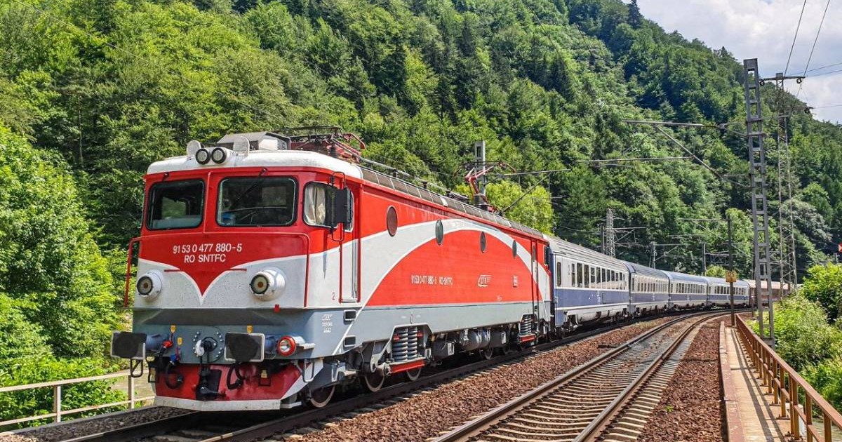 Un tren de călători cu locomotivă roșie merge pe calea ferată din România într-o zonă montană, este înconjurată linia de cale ferată de arbori cu frunze verzi.