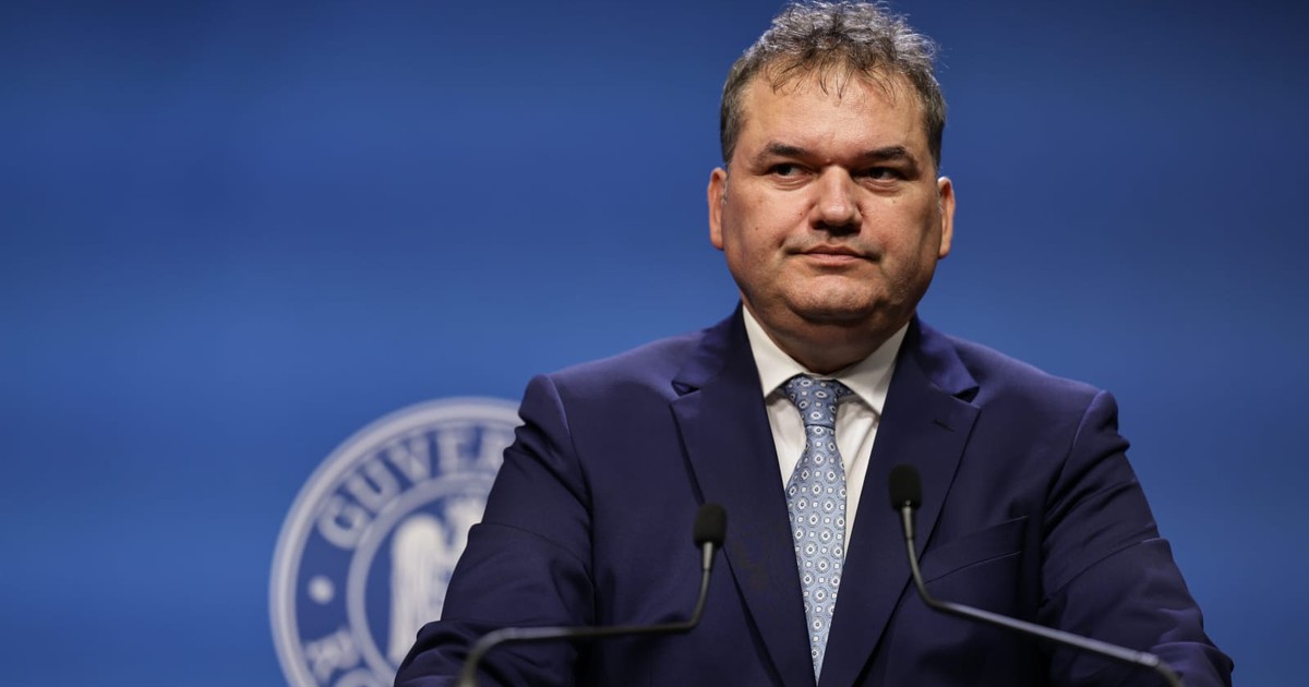 Cseke Attila, îmbrăcat într-un sacou bleumarin, o cămașă albă și o cravată gri, stând în fața unui pupitru cu două microfoane, cu emblema guvernului româniei în spate