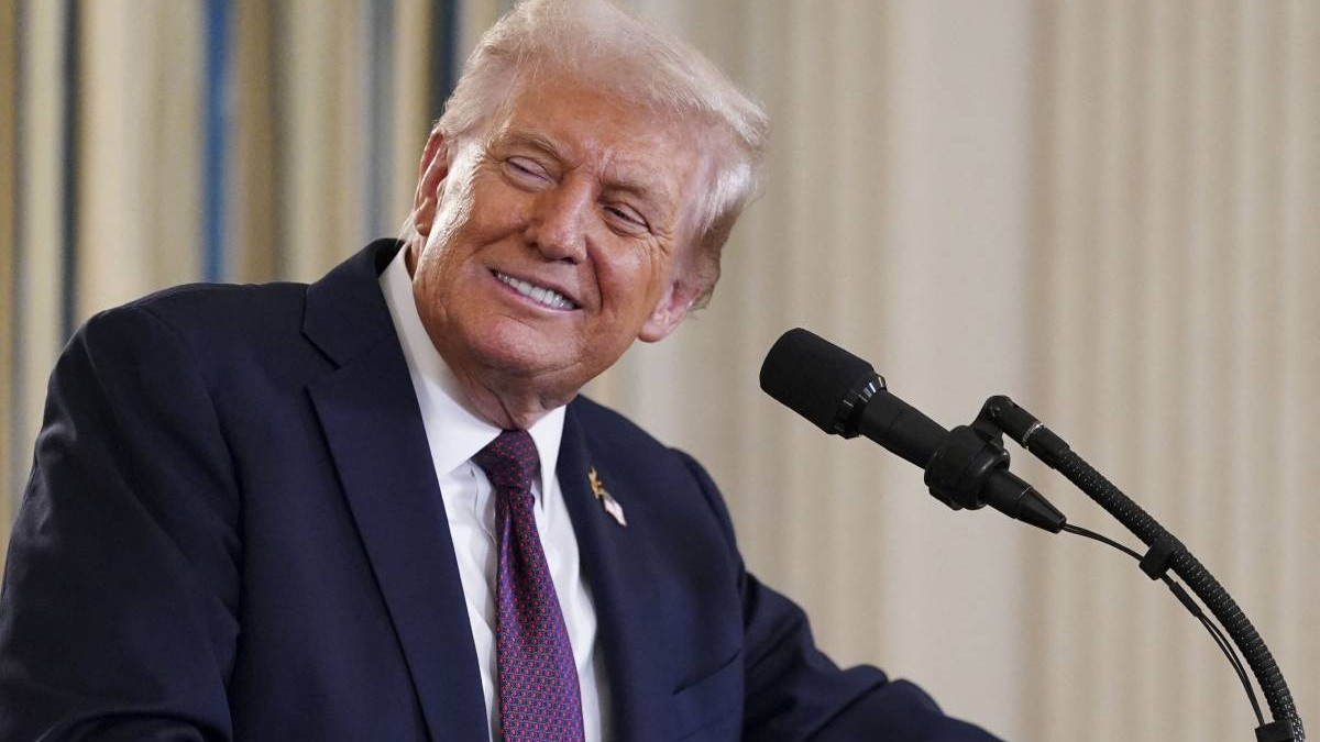 Președintele american Donald Trump, în timpul unei conferințe de presă la Casa Albă, poartă un costum negru și o cravată violet și zâmbește în fața camerei