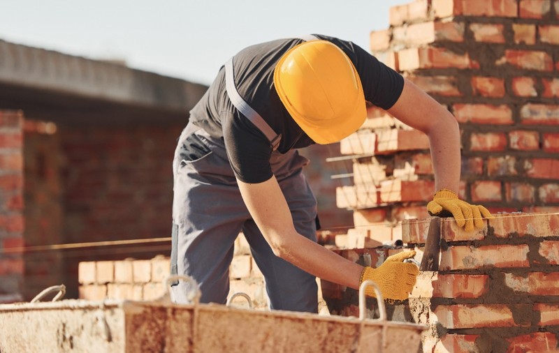 Un muncitor în construcții care poartă cască galbenă de protecție și mănuși, lucrând la zidirea unui perete din cărămidă roșie pe un șantier.