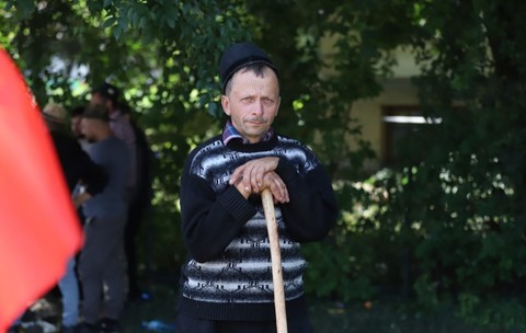 Ciobanul Liviu din Bihor, un bărbat cu un pulover albastru de lână, lângă un gard viu din București. Își ține capul sprijinit într-un toiag