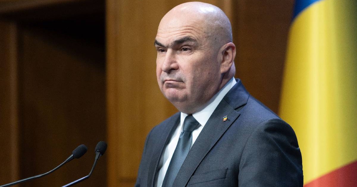 Ilie Bolojan, îmbrăcat într-un costum, cu steagul românesc pe fundal, vorbește în timp ce schițează o grimasă