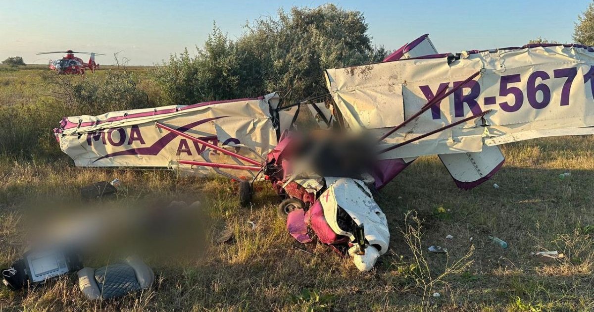 un avion alb de mici dimensiuni, cu detalii roz, prăbușit la pământ, cu aripile deteriorate. lânga epava avionului se pot observa scaunele avionului, rupte și aruncate la o distanța de un metru
