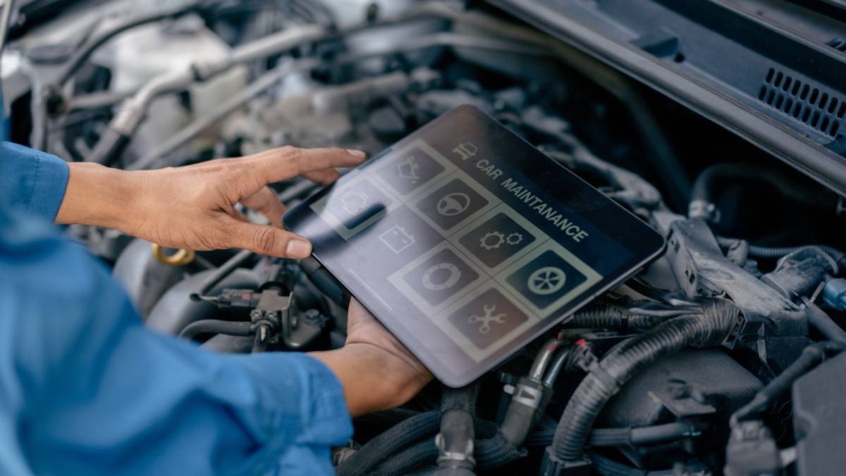 Mecanic auto, îmbrăcat cu o salopetă albastră, în timp ce face inspecția unei mașini. Ține în mână o tabletă, deasupra motorului unei mașini.