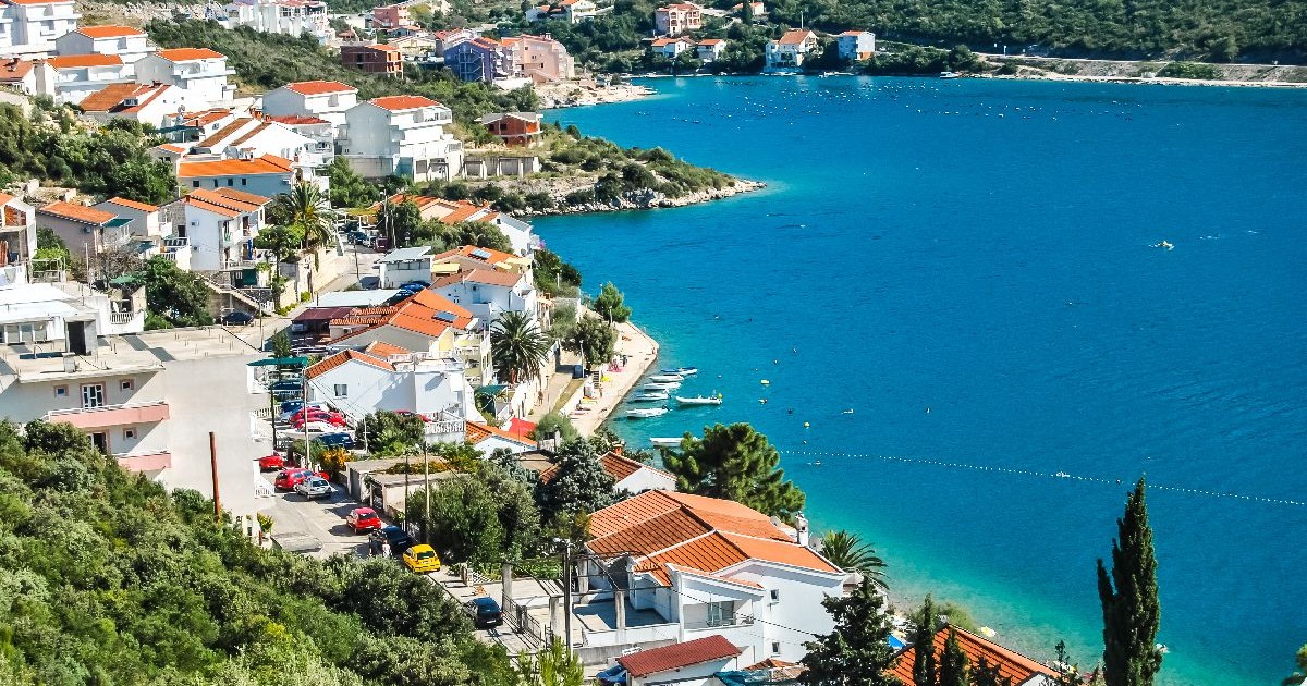 Imaginea prezintă o vedere panoramică a unei localități de coastă, cu numeroase case albe cu acoperișuri roșii dispuse în trepte pe un versant împădurit ce coboară spre apele turcoaz ale unui golf liniștit.