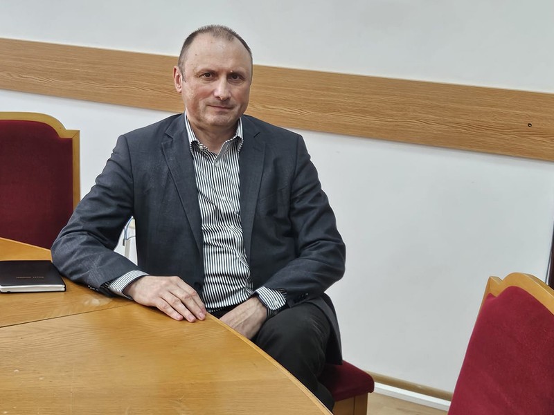 Un barbat in costum gri sta la o masa si acordă un interviu
