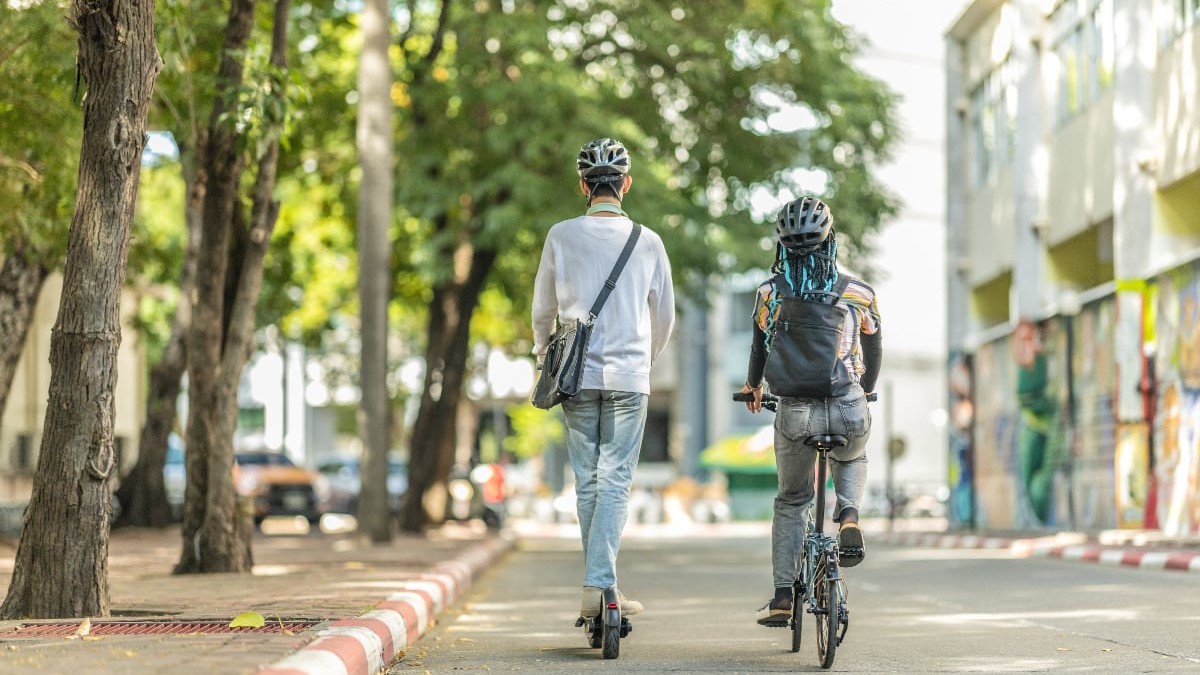 Imaginea surprinde doi tineri care se deplasează pe o stradă liniștită, umbrită de copaci – unul pe trotinetă electrică, celălalt pe bicicletă. Amândoi poartă căști de protecție și rucsacuri, sugerând fie o deplasare urbană obișnuită, fie o ieșire recreativă.