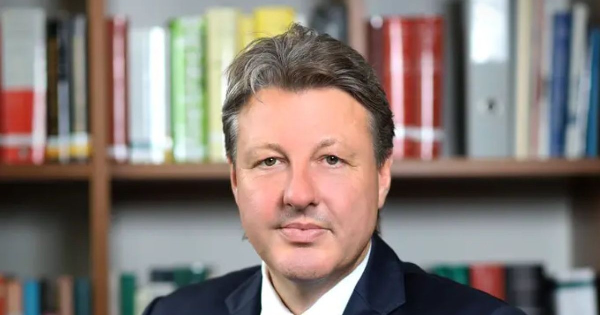 Dr. Aleksander Ospelt, îmbrăcat în costum, iar pe fundal o bibliotecă cu cărți