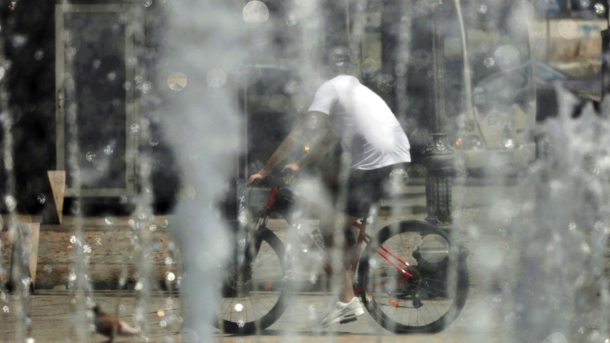 Un tânăr pe bicicletă, fotografiat cu spatele, în tricou alb și cu cască pe cap, trece prin dreptul unor fântâni din București, într-o zi caniculară de vară, București, 17 iulie 2025.